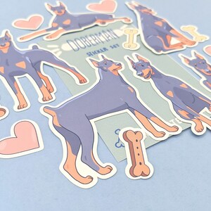 May include: A set of Doberman Pinscher dog stickers. The stickers are in various poses, including sitting, standing, and running. The stickers are on a light blue background with a white border. The stickers are part of a sticker set called "Doberman Sticker Set".