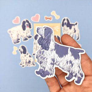 May include: A collection of stickers featuring a blue and white Cocker Spaniel dog illustration. The stickers include various poses of the dog, along with heart and bone-shaped designs. The dog stickers are held in a hand against a light blue background.