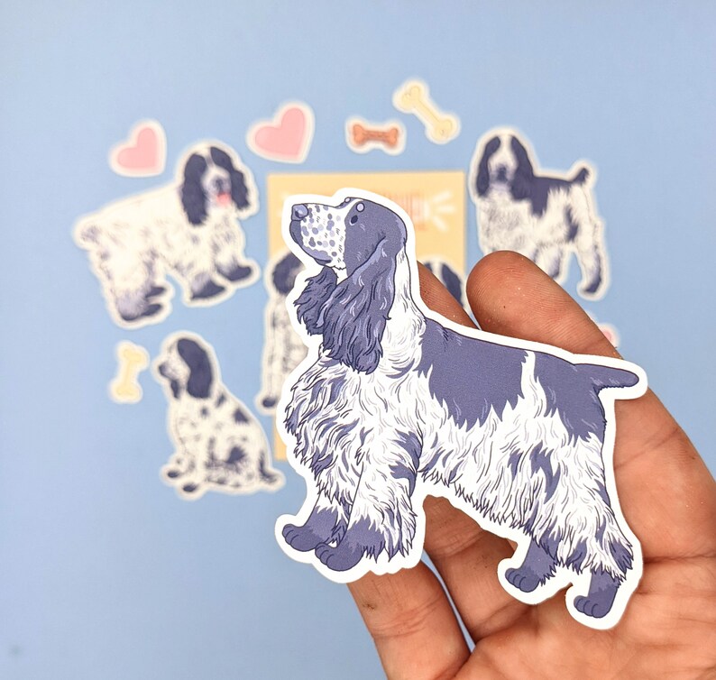 May include: A collection of stickers featuring a blue and white Cocker Spaniel dog illustration. The stickers include various poses of the dog, along with heart and bone-shaped designs. The dog stickers are held in a hand against a light blue background.
