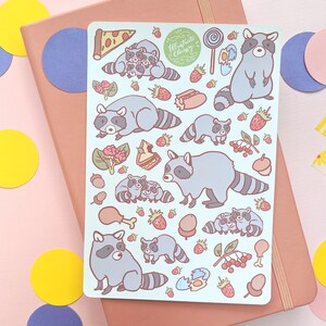 May include: A light blue sticker sheet featuring cartoon raccoons, pizza, strawberries, and other food items. The sticker sheet has the text "Illustrate & Chrissy" in a green circle. The sheet is on a pink notebook.