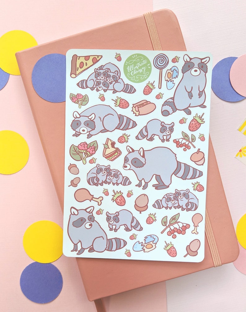 May include: A light blue sticker sheet featuring cartoon raccoons, pizza, strawberries, and other food items. The sticker sheet has the text "Illustrate & Chrissy" in a green circle. The sheet is on a pink notebook.