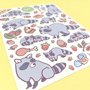 May include: A sticker sheet featuring cartoon raccoons and various food items. The raccoons are depicted in different poses, with gray and white fur. Food illustrations include pizza, strawberries, and a drumstick. The background is a light yellow.