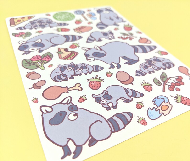 May include: A sticker sheet featuring cartoon raccoons and various food items. The raccoons are depicted in different poses, with gray and white fur. Food illustrations include pizza, strawberries, and a drumstick. The background is a light yellow.
