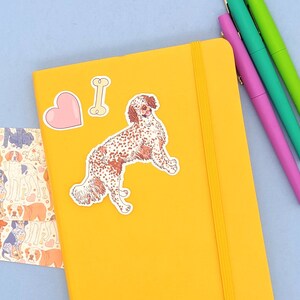 Pu&ograve; includere: Un quaderno giallo con un adesivo di un cane, un adesivo a forma di cuore e un adesivo a forma di osso. Il quaderno ha una chiusura con elastico giallo.