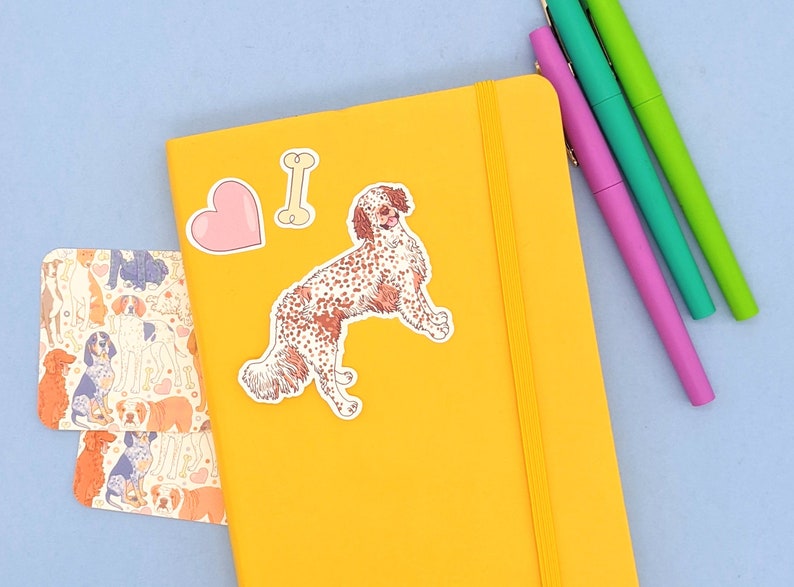 Pu&ograve; includere: Un quaderno giallo con un adesivo di un cane, un adesivo a forma di cuore e un adesivo a forma di osso. Il quaderno ha una chiusura con elastico giallo.