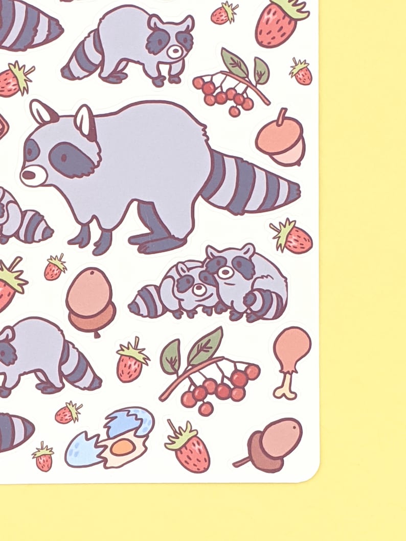 May include: A sheet of stickers featuring cartoon raccoons in various poses, along with strawberries, acorns, and berries. The raccoons are primarily gray with black stripes and accents. The strawberries are red, and the acorns are brown.