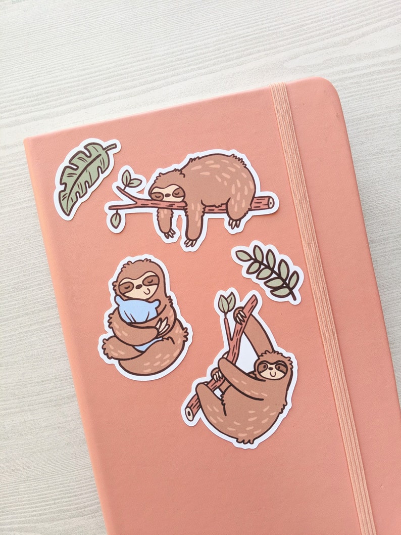 May include: Three brown sloth stickers on a pink background. One sloth is sleeping on a branch, another is holding a blue pillow, and the third is hanging from a branch.