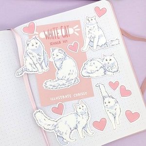 May include: A set of white cat stickers with pink hearts and a pink card that reads "WHITE CAT STICKER SET" and "ILLUSTRATE CHRISSY" on a white dotted notebook. Pens are visible in the background.
