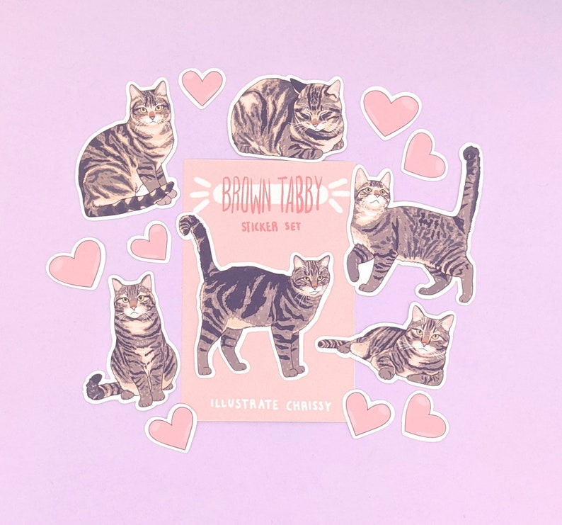 May include: A set of brown tabby cat stickers with pink hearts. The stickers feature different poses of the cats, including sitting, lying down, and walking. The text "Brown Tabby Sticker Set" is printed on a white background.