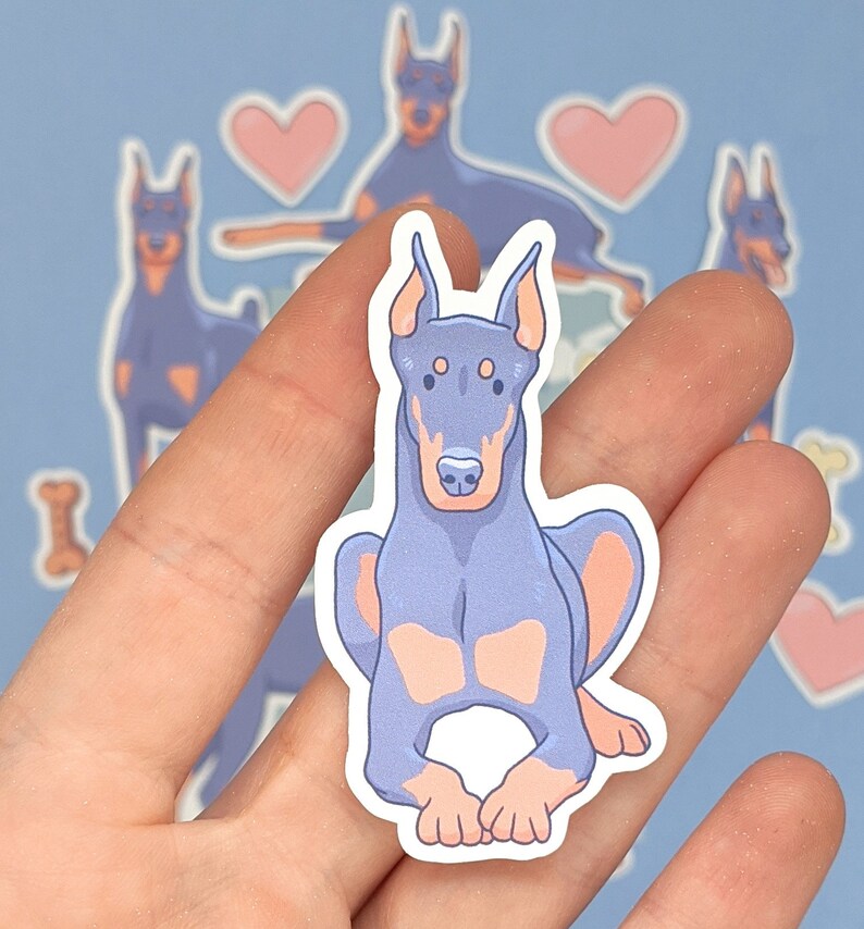 May include: A sticker featuring a cartoon illustration of a blue and pink Doberman Pinscher dog. The dog is sitting with its legs crossed and has a friendly expression.