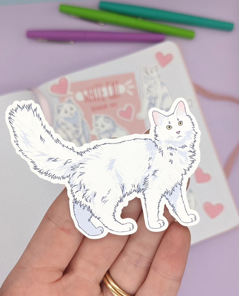 May include: A white cat sticker with a fluffy tail and blue accents. The sticker is held in a hand, with a notebook and pens in the background. The notebook has cat illustrations and the text "WHITE CAT". Pink hearts are also visible.