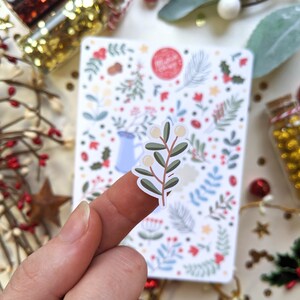 May include: A white sticker with a green branch with white berries. The sticker is on a finger and the background is a sheet of paper with a Christmas themed pattern.