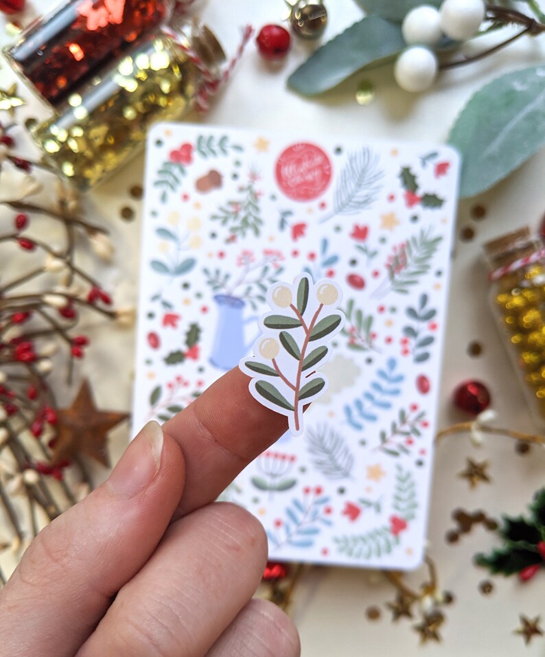 May include: A white sticker with a green branch with white berries. The sticker is on a finger and the background is a sheet of paper with a Christmas themed pattern.