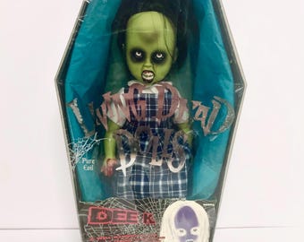 Puede incluir: Una muñeca Living Dead Dolls en una caja con forma de ataúd. La muñeca tiene piel verde, pelo negro y un vestido a cuadros. La caja tiene las palabras "I'm Dead" y "Pure Evil".