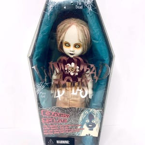 May include: A doll in a coffin-shaped box. The doll has pale skin, yellow eyes, and brown hair. It wears a brown top with a flower and the words "I'm Dead". The box has the words "Pure Evil" and "Daisy Slab" on it.