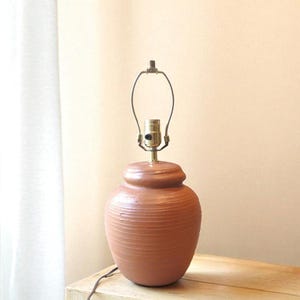 May include: A terracotta-colored ceramic table lamp with a textured, rounded base and a brass-colored lamp holder. The lamp sits on a wooden surface, with a white curtain in the background.