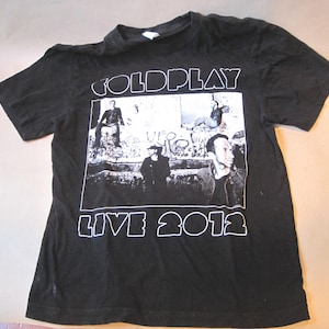 May include: A black short-sleeve t-shirt featuring a white graphic print on the front. The design includes the text COLDPLAY at the top and LIVE 2012 at the bottom, surrounding a black and white photograph of band members in a graffiti-covered area.