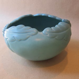 May include: A light blue ceramic bowl with a scalloped rim and leaf-like designs. The bowl has a rounded shape and a smooth, matte finish. The interior of the bowl is a darker shade of blue.