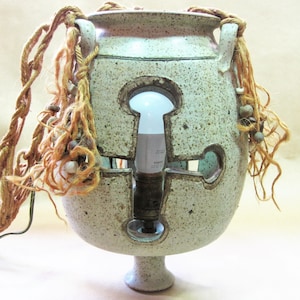 May include: A speckled ceramic lamp with a keyhole design, featuring a light bulb inside. The lamp has rope and decorative elements attached to the sides. The lamp is a unique home decor item.