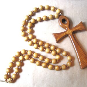 May include: A necklace featuring a large, polished wooden ankh pendant with a visible grain pattern, connected to a long strand of round, light brown wooden beads separated by small, metallic copper-colored spacer beads throughout the design.