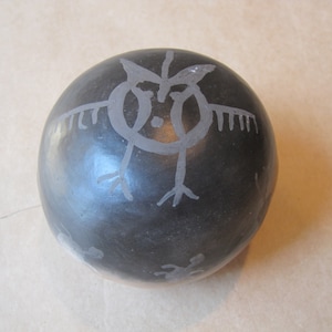 May include: A spherical black pottery vessel with a smooth, polished finish. The surface features light gray, hand-etched line drawings of a stylized bird-like figure with outstretched wings and smaller, indistinct animal shapes near the base.