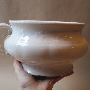May include: A white ceramic chamber pot with a handle. The pot has a wide, rounded body and a small, decorative foot. The handle is curved and ornate. The pot is held by a hand.