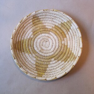 May include: A round, shallow woven basket featuring a coiled design with alternating sections of natural cream and muted olive green fibers. The basket has a spiral pattern radiating from the center toward the reinforced, stitched outer rim edge.
