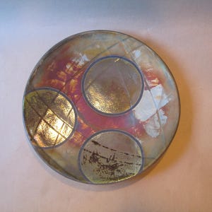 May include: A round ceramic plate featuring a multi-colored abstract design with shades of red, yellow, and white. The surface is decorated with three distinct circular motifs, each containing a grid-like pattern in metallic gold tones.