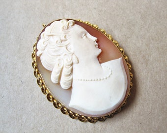 May include: A cameo brooch featuring a carved portrait of a woman with a detailed hairstyle, set in a gold-toned oval frame.