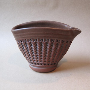 May include: A brown ceramic bowl with a textured, geometric pattern around the exterior. The bowl has a spout and a wide opening. The bowl is approximately 6 inches wide and 4 inches tall.