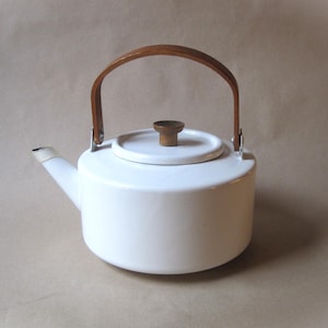 May include: A white enamel teapot featuring a cylindrical body, a short spout, and a removable lid with a small brown wooden knob. The teapot includes a curved, arched wooden handle attached to the sides with silver-toned metal hardware.