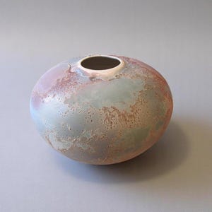 May include: A round ceramic vase with a mottled glaze in shades of blue, pink, and brown. The vase has a smooth, rounded shape and a small opening at the top.