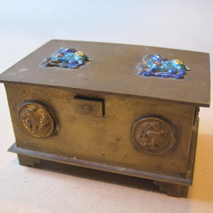 May include: A small, rectangular brass box with a hinged lid. The lid is decorated with two clusters of blue and gold enamel. The box has four small feet and two round brass plaques on the front.