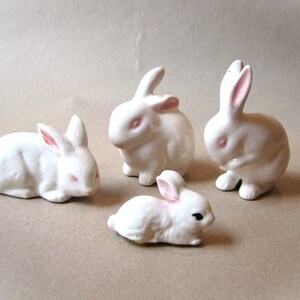 May include: Four white ceramic bunny figurines. The bunnies are in various poses, including sitting, lying down, and standing. The bunnies have pink noses and eyes.
