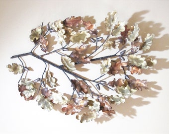 May include: Metal wall art depicting a branch with leaves and acorns. The leaves are in shades of white, brown, and copper. The branch is dark gray. This decorative piece adds a touch of nature to any space.