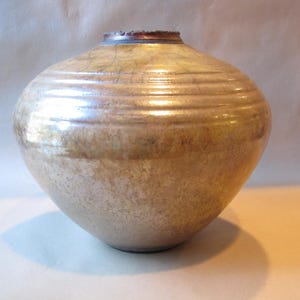 May include: A large, round, ceramic vase with a light brown glaze. The vase has a slightly iridescent finish and a subtle texture.