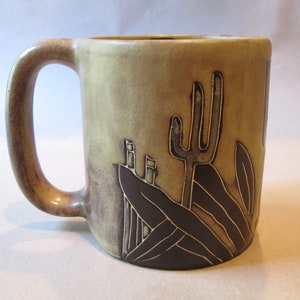 May include: A brown ceramic mug with a handle. The mug has a desert scene design with a cactus, mountains, and plants in brown and tan.