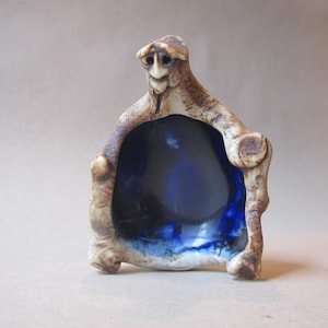 May include: A handcrafted ceramic sculpture featuring a stylized figure with a smiling face and a deep blue interior. The sculpture has a textured, earthy appearance with brown and beige tones, and is likely a decorative art piece.