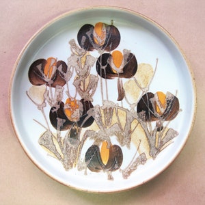May include: A round, white ceramic plate with a floral design. The flowers are brown, orange, and beige, with a stylized, abstract appearance. The plate has a light brown rim and is suitable for serving food.