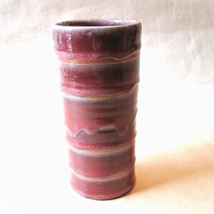 May include: A tall, cylindrical ceramic vase with a red and white glaze. The vase has a banded design with a slightly uneven, drippy glaze.