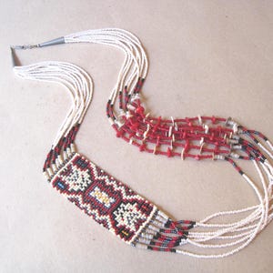 May include: A beaded necklace with a geometric design. The necklace features a rectangular pendant with a pattern of red, black, white, and blue beads. The necklace has multiple strands of white beads and red beaded sections.