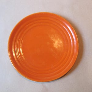 May include: A round, vibrant orange ceramic plate featuring a smooth, glossy finish and a series of three concentric circular ridges embossed along the outer rim, creating a distinct textured pattern on the surface of the dinnerware piece.