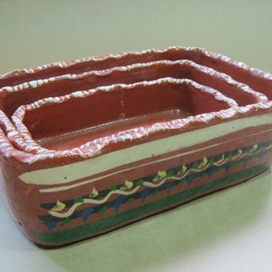 May include: A set of three nested rectangular terracotta baking dishes with a hand-painted exterior featuring horizontal bands of white, green, and blue, topped with a wavy line pattern and a textured, uneven rim finish in white and red.