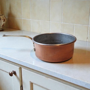 May include: A copper saucepan with a long, brass handle. The pot has a rounded shape and a hammered texture. It sits on a white countertop in a kitchen setting. A decorative vase is in the background.