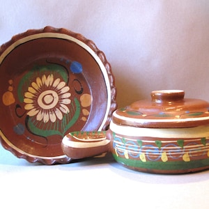 May include: A set of two hand-painted terracotta pottery pieces. The larger piece is a covered casserole dish with a handle. The smaller piece is a shallow dish with a scalloped edge. Both pieces feature floral designs in shades of brown, green, yellow, and blue.