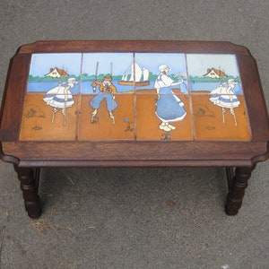 May include: A wooden coffee table with a tiled top featuring a colorful illustration of people on stilts. The tiles are arranged in a grid pattern and depict a scene of people walking on stilts by a body of water.