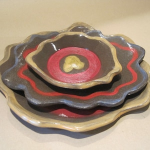 May include: A set of three ceramic bowls, each with a unique design. The largest bowl is at the bottom, followed by two smaller bowls stacked on top. The bowls have a dark brown interior with red and blue accents, and a gold-colored rim.