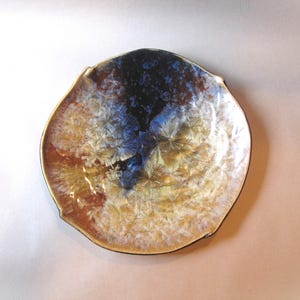 May include: A decorative ceramic plate with a unique, organic shape. The plate features a blend of colors, including brown, blue, and white, with a crystalline pattern. The rim is a dark brown color. The plate is approximately 8 inches in diameter.