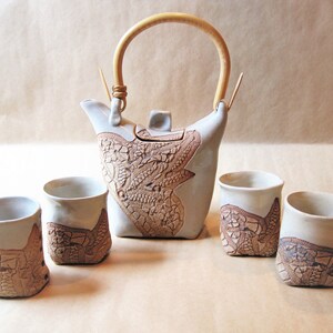 May include: A ceramic tea set featuring a teapot and four cups. The teapot has a light beige body with a textured, brown floral design and a bamboo handle. The cups have a similar design. The set is ideal for serving tea.