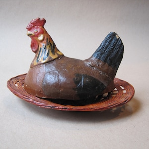May include: A ceramic rooster figurine with brown, black, and yellow paint, sitting on a woven wicker nest.